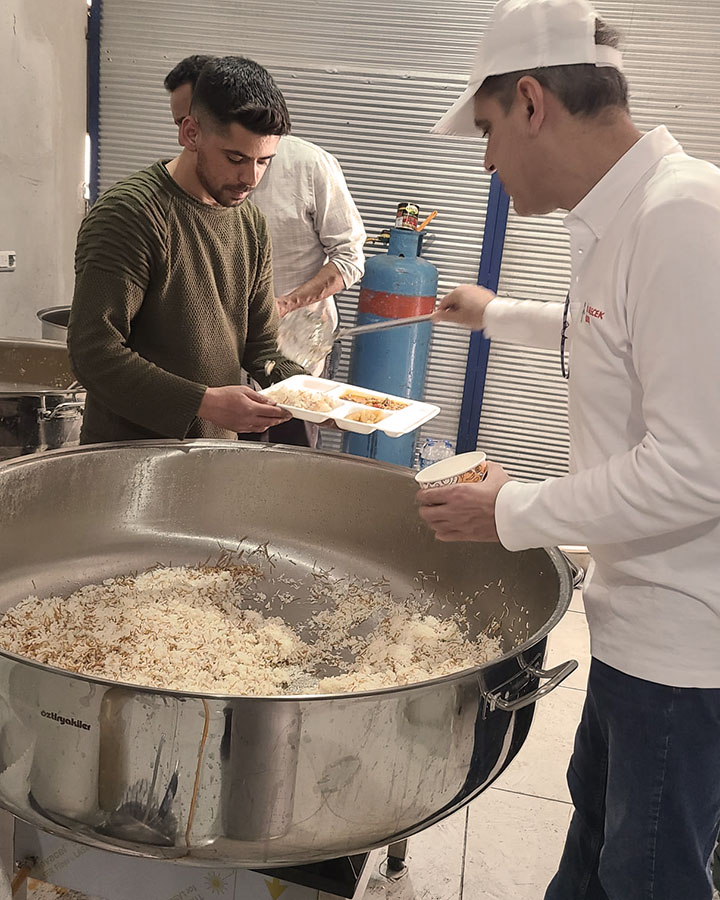 A male in white hat and tshirt serving another young male rice from a large pot at a relief location in Turkey